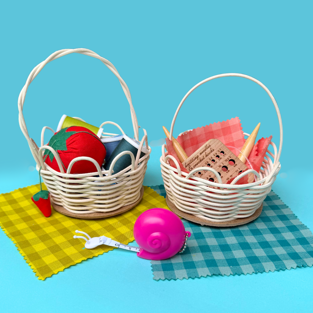 Two mini rattan baskets filled with sewing and knitting notions, sitting on top of yellow and blue gingham fabric swtaches. A pink snail tape measure sits in front of the baskets.